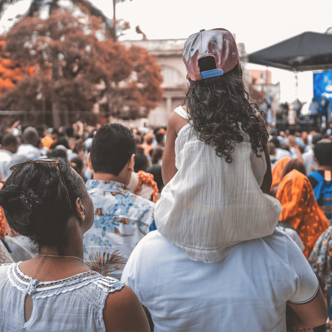 View from behind of a little girl sitting on a man's shoulders in a crowd attending a concert.