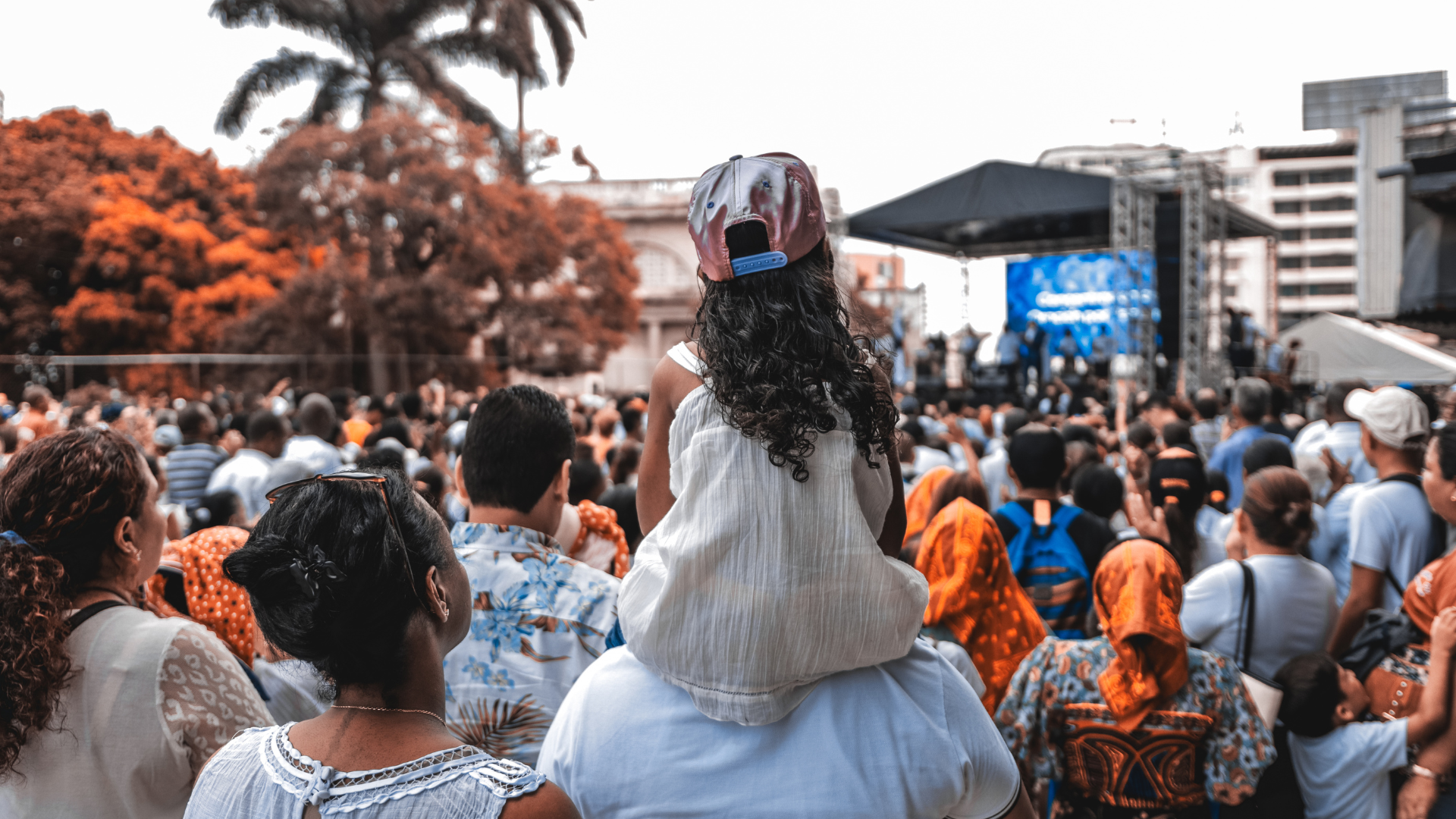 View from behind of a little girl sitting on a man's shoulders in a crowd attending a concert.